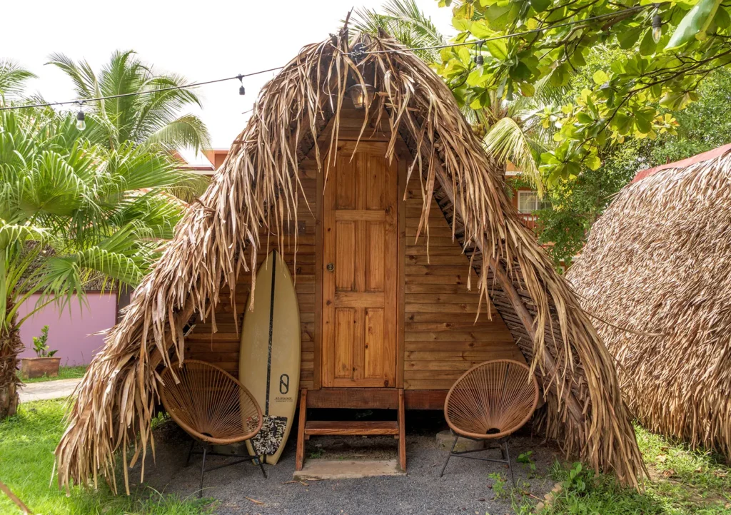 A thatched A-frame tepee with a surfboard by the door and two chairs outside, among palms